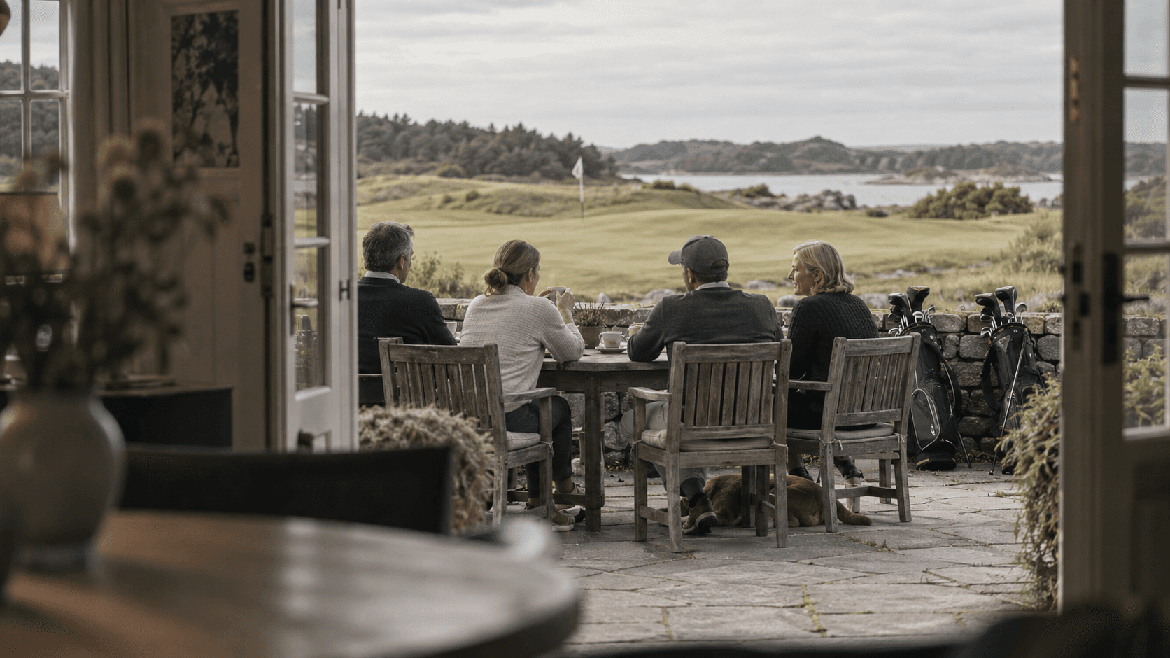 Four members sit over coffee on the clubhouse terrace after their round, bags against the wall and the course beyond.