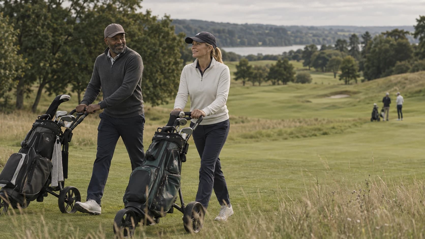 Two golfers push their trolleys up the fairway, talking, with the lake and the next group behind them.
